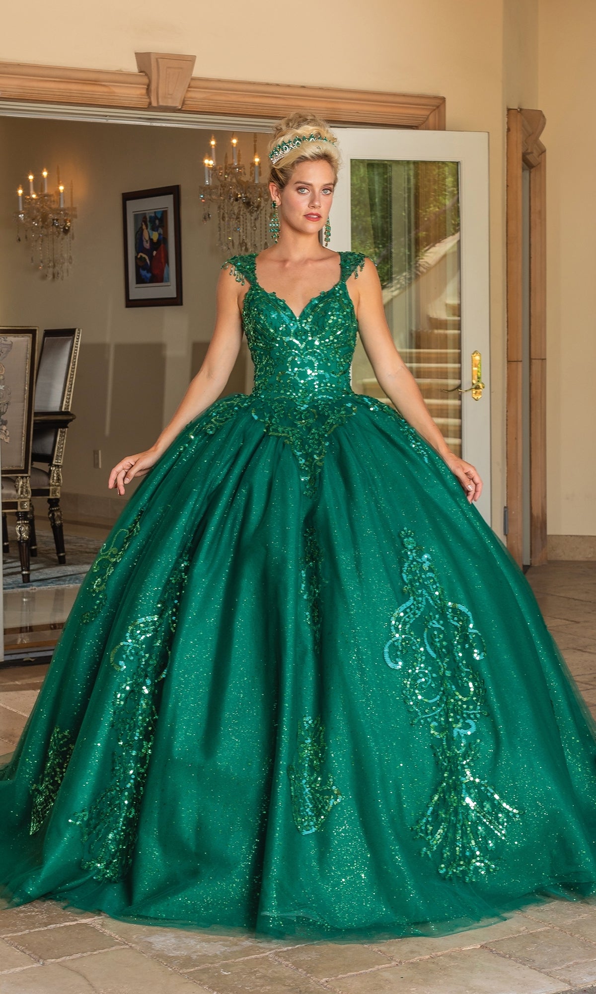 A woman stands indoors wearing the Quinceanera Dress 1758 by Dancing Queen, an emerald green A-line ball gown with cap sleeves, a voluminous skirt, glitter lace embroidery, and intricate detailing.